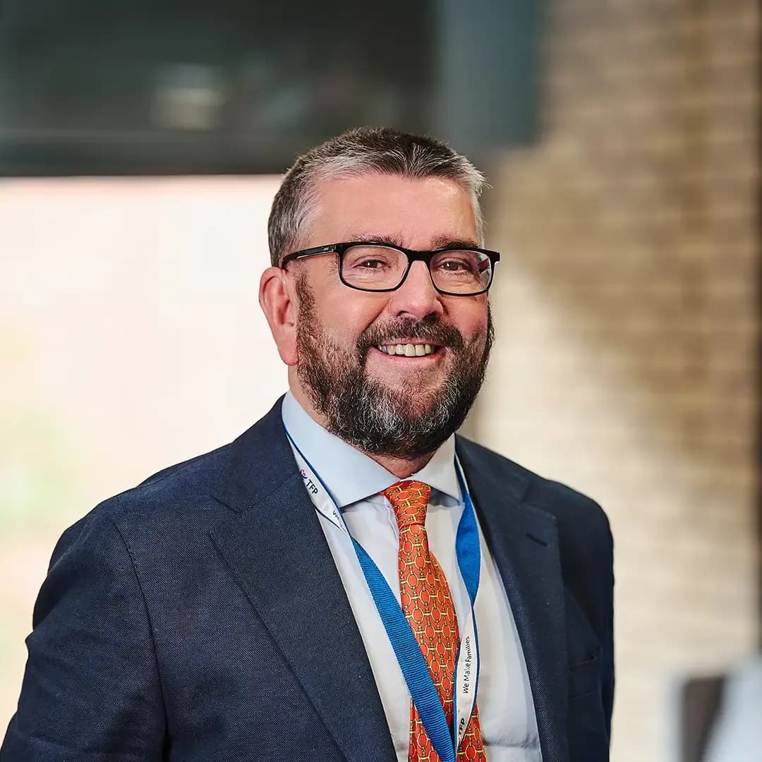 Man with glasses and a beard wearing a suit, orange tie, and lanyard smiles in a professional indoor setting.