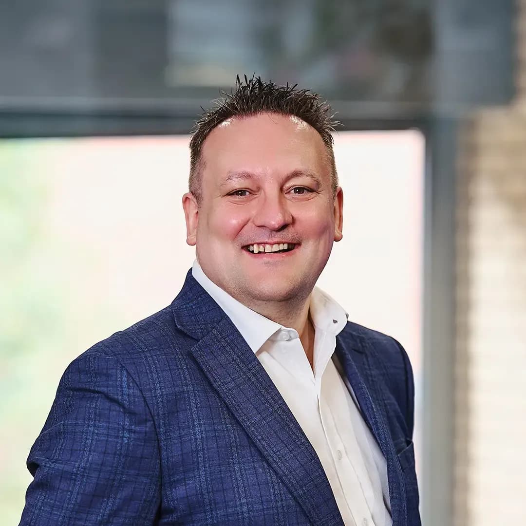Smiling man in a blue suit and white shirt, standing in a well-lit room with a blurred background.