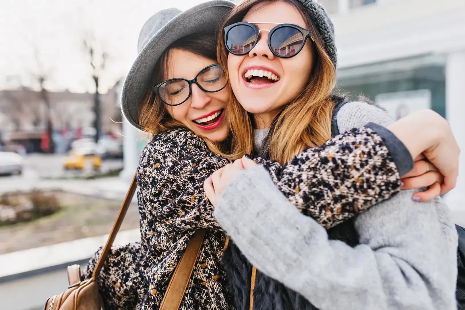Two women in winter clothing, wearing sunglasses and hats, joyfully hugging and smiling outdoors on a sunny day.