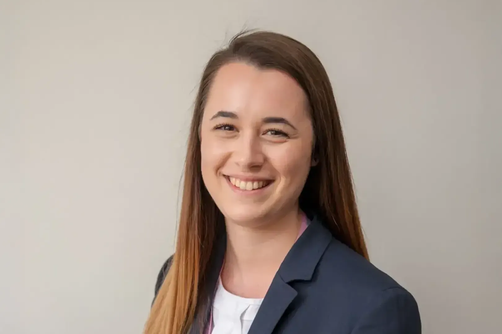 Smiling person with long brown hair wearing a navy blazer and white shirt against a plain background.