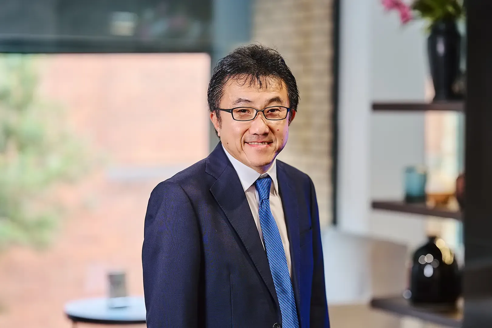 Man in a suit and blue tie smiling, standing indoors with a blurred background of a window and shelves.